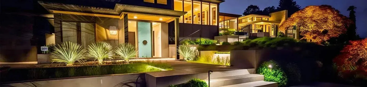Modern house exterior at night with warm wall lights and garden path illumination highlighting plants and concrete steps