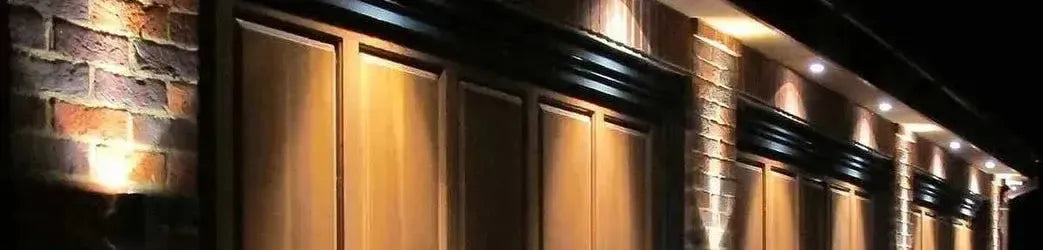Round silver downlights recessed into a dark eaves soffit casting warm light onto a brick wall and timber garage doors