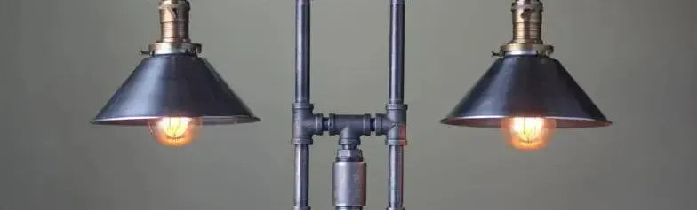 Industrial floor lamp with twin conical metal shades and brass fittings on a dark grey pipe frame against a neutral wall