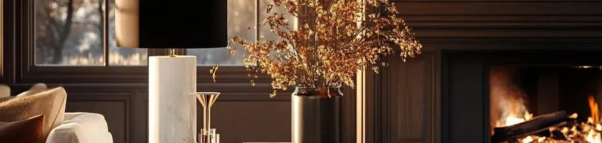 White marble cylindrical table lamp with a black shade beside a vase on a table in a cosy room with a fireplace