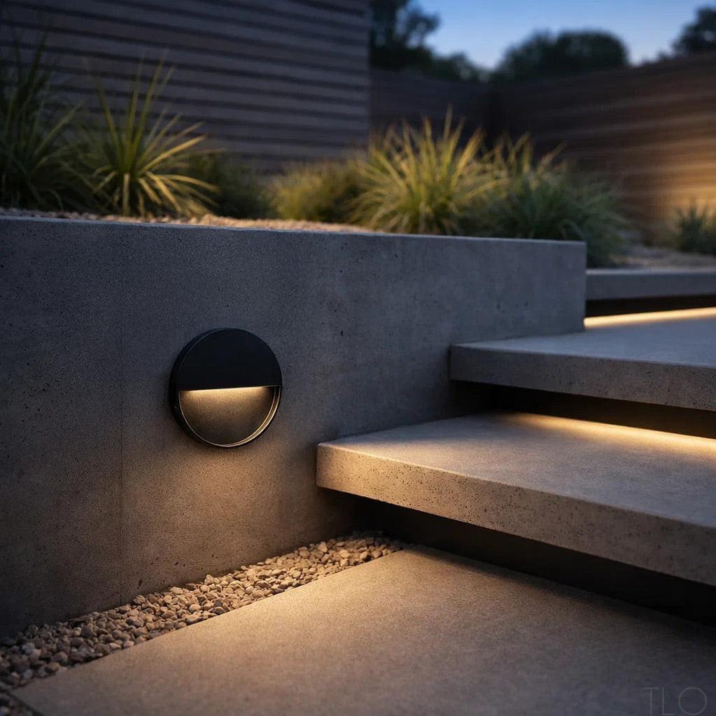 Round black aluminium LED step light mounted on a concrete garden wall, illuminating outdoor stairs at dusk.