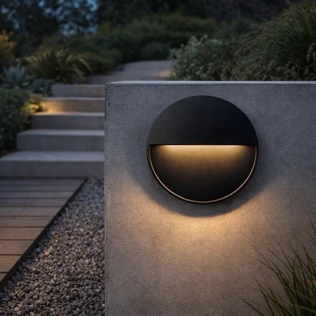 Round black aluminium LED outdoor step light mounted on a concrete garden wall, emitting a warm downward glow.