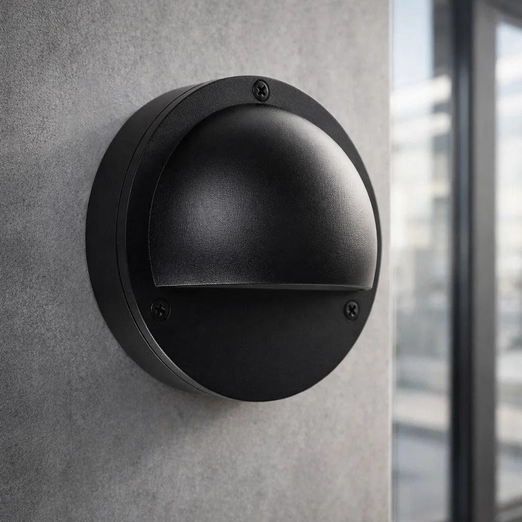 Round matt black aluminium Pinta step light with an eyelid design mounted on a grey exterior wall.